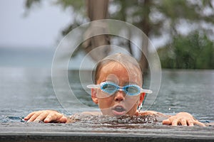 Cute little caucasian blue-eyed boy looking mindfully into camera through swim goggles after diving in pool