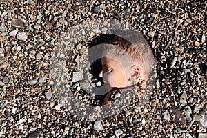 Cute little caucasian boy lying dug under stone pebble on summer sunny sea beach sunbathing