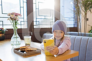 Cute little caucasian girl with blonde hair in trendy casual clothes using mobile phone in modern cafe at spring. Video