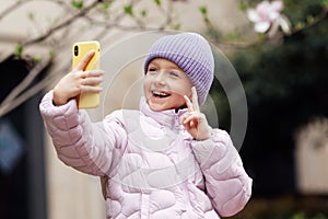 Cute little caucasian girl with blonde hair in trendy casual clothes using mobile phone outdoor at spring. Video chat