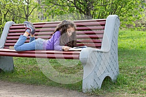 Cute little caucasian girl reading book lying on a bench in spring park