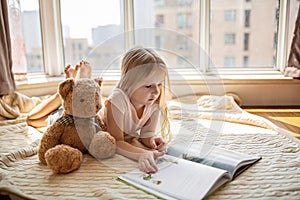 Cute little caucasian girl reading a book with stuffed teddy bear toy. Stay at home during coronavirus covid-19 pandemic