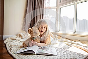 Cute little caucasian girl reading a book with stuffed teddy bear toy. Stay at home during coronavirus covid-19 pandemic