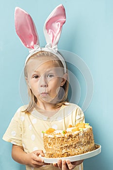 Cute little caucasian girl wearing bunny ears and holding delicious carrot cake on the blue background