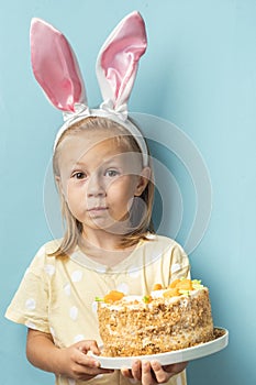 Cute little caucasian girl wearing bunny ears and holding delicious carrot cake on the blue background. Happy Easter