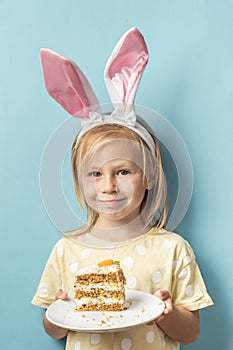 Cute little caucasian girl wearing bunny ears and holding delicious carrot cake on the blue background. Happy Easter