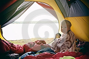 Cute little child boy sitting in camping tent, wrapped in wool blanket. Family weekend outdoor, local travel on nature, trekking,