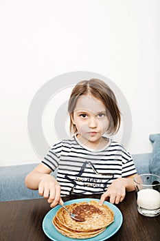 Cute little child eating homemade pancakes with chocolate cream and milk at home