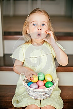 Cute little child girl with blonde hair on Easter day. Girl holding basket with painted eggs and sitting on the stairs at home