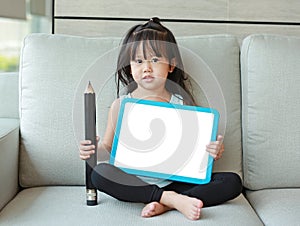 Cute little child girl holding empty white blackboard with big pencil sitting on fabric sofa in library room. Education concept