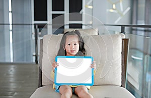 Cute little child girl holding empty white blackboard on fabric sofa in library room