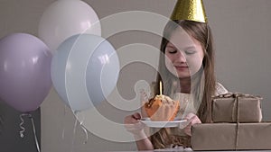 Cute little child girl in party hat sitting surrounded by air balloons and gifts going to celebrate birthday party