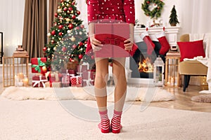 Cute little child hiding gift box behind her back in room with Christmas tree, closeup