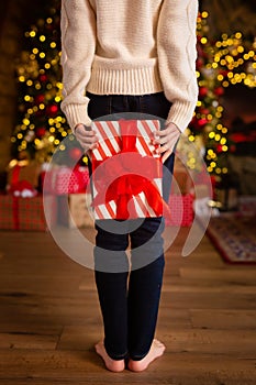 Cute little child hiding gift box behind her back in room with Christmas tree, closeup