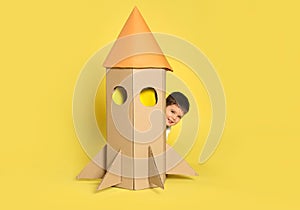 Cute little child playing with cardboard rocket on yellow background
