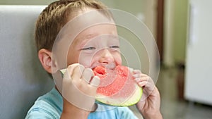 A cute little child is sitting at the kitchen table and is eating a juicy watermelon with an appetite. Face laughing boy