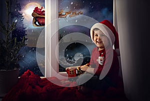 Cute little child on window sill at home waiting for Santa Claus. Christmas celebration