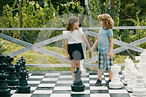 Cute little children playing on big chess board. Portrait of two happy young kids play chess at the summer park.