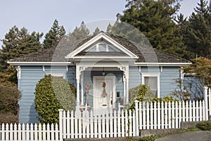 Cute little cottage with blue siding and victorian touches and wood shingles and white picket fence against tall pine trees