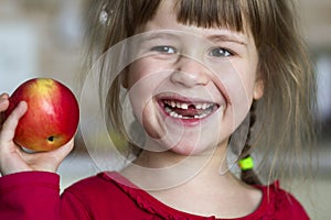 A cute little curly toothless girl smiles and holds a red apple. Portrait of a happy baby eating a red apple. The child loses milk