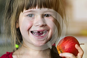 A cute little curly toothless girl smiles and holds a red apple. Portrait of a happy baby eating a red apple. The child loses milk