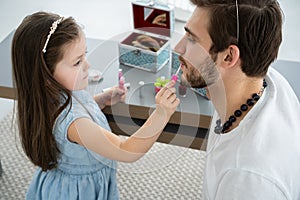 Cute little daughter and her handsome young dad in crowns are playing together in child`s room. Girl is doing her dad a
