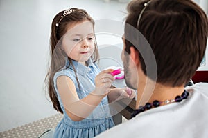 Cute little daughter and her handsome young dad in crowns are playing together in child`s room. Girl is doing her dad a