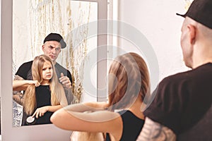 Cute little daughter and her tattoed dad are playing together near a mirror. Dad is doing his daughter`s hair. Family