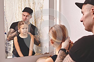 Cute little daughter and her tattoed dad are playing together near a mirror. Dad is doing his daughter`s hair. Family