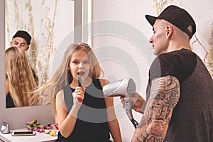 Cute little daughter and her tattoed dad are playing together near a mirror. Dad is doing his daughter`s hair. Family