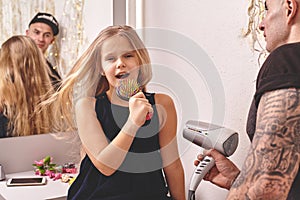 Cute little daughter and her tattoed dad are playing together near a mirror. Dad is doing his daughter`s hair. Family