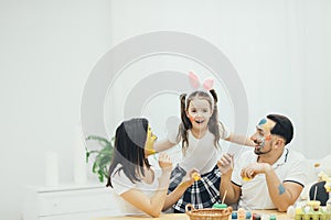 Cute little daughter with pigtails and bunny ears is hugging her parents. Mother and father painting easter eggs