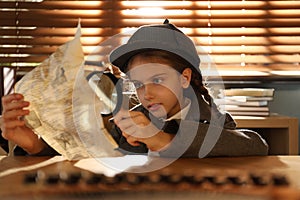 Cute little detective exploring document with magnifying glass at table in office