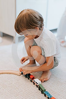 Cute little five year old kid boy in a white t-shirt playing with a wooden railway and toy trains on the floor on the carpet in th