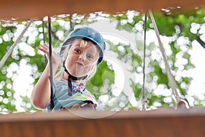 Cute little funny caucasion blond girl in helmet having fun walking by rope suspension bridge in adventure park