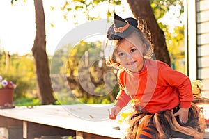 A cute little girl 2-3 in an orange and black witch costume sits next to pumpkins on the terrace of a wooden gray house