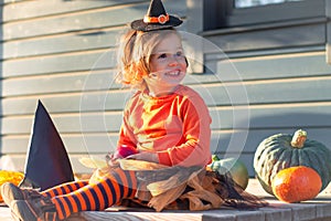 A cute little girl 2-3 in an orange and black witch costume sits next to pumpkins on the terrace of a wooden gray house