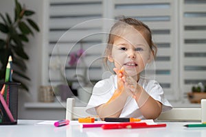 Cute little girl 2-4 in a white t-shirt draws at home with colorful felt-tip pens and pencils