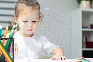 Cute little girl 2-4 in a white t-shirt draws at home with colorful felt-tip pens and pencils