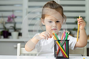 Cute little girl 2-4 in a white t-shirt draws at home with colorful felt-tip pens and pencils