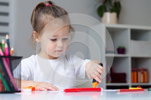 Cute little girl 2-4 in a white t-shirt draws at home with colorful felt-tip pens and pencils