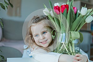 Cute little girl of 3 years old in a cozy knitted sweater hugs a vase with a bouquet of white and pink tulips in a home interior.