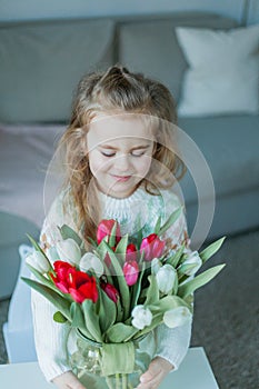 Cute little girl of 3 years old in a cozy knitted sweater hugs a vase with a bouquet of white and pink tulips in a home interior.