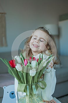 Cute little girl of 3 years old in a cozy knitted sweater hugs a vase with a bouquet of white and pink tulips in a home interior.