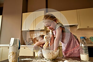 Cute little girl in apron kneading dough while making cookies together with her brother on the kitchen table at home