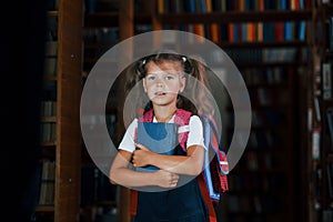 Cute little girl with backpack stands in the library full of books. Conception of education