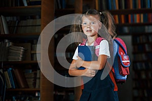 Cute little girl with backpack stands in the library full of books. Conception of education