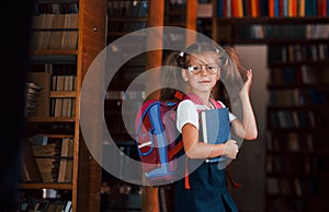 Cute little girl with backpack stands in the library full of books. Conception of education