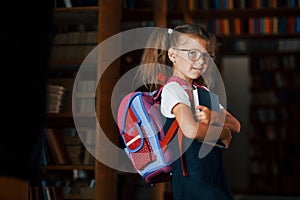 Cute little girl with backpack stands in the library full of books. Conception of education