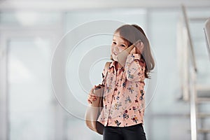 Cute little girl with bag indoors in the office or airport talking by phone